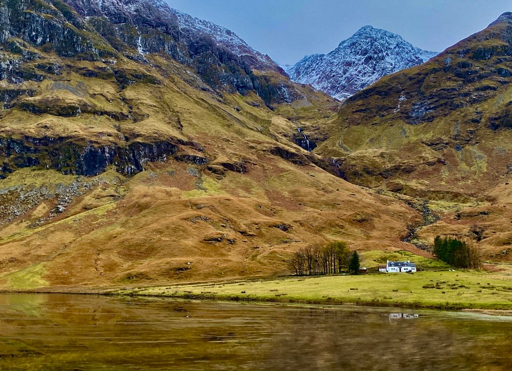 Snow capped Highland mountains and a lone cottage beside a loch in Glencoe, Scotland