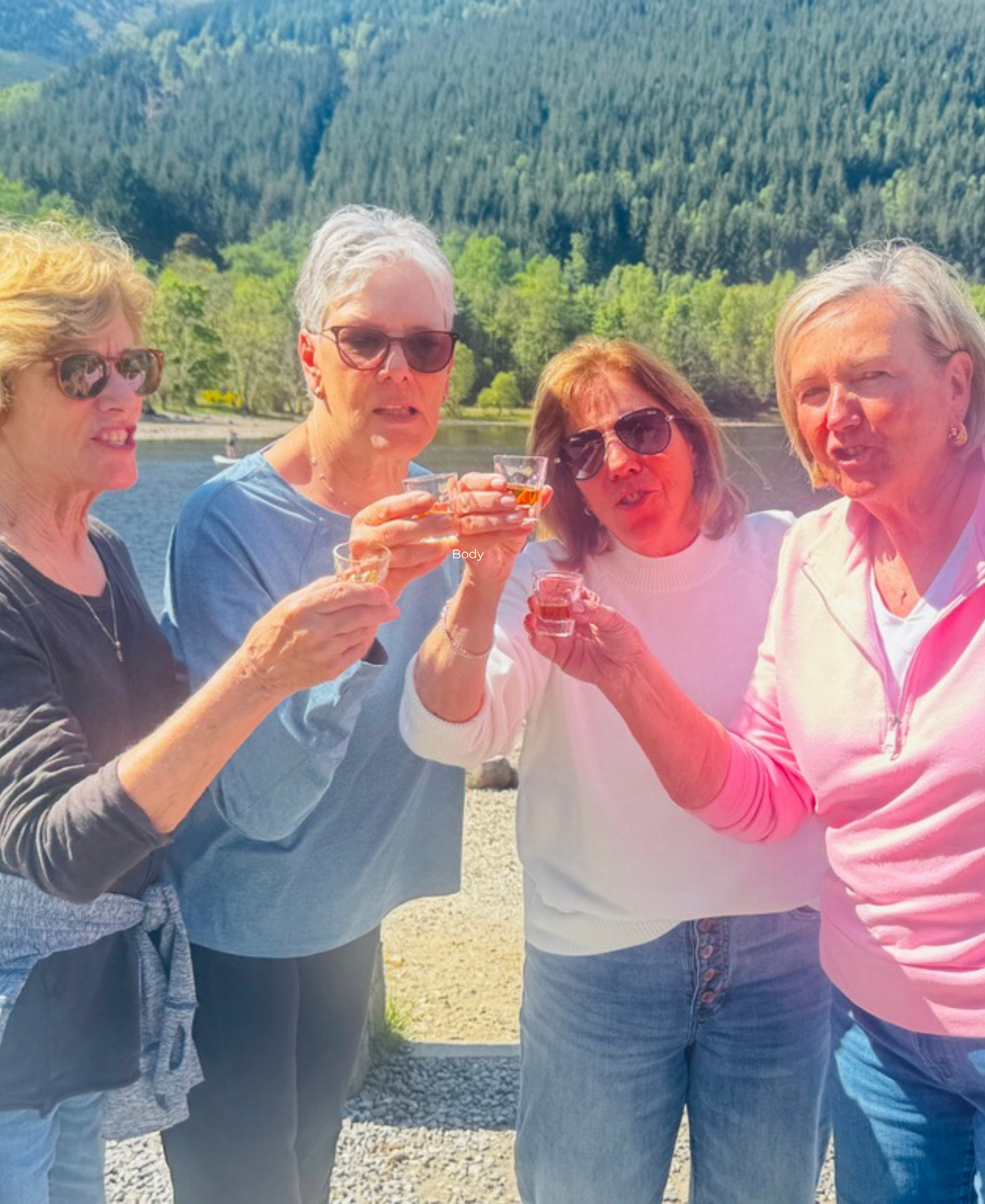 A Toast by the Loch Four women toasting with small glasses of whisky beside a Scottish loch surrounded by forested hills