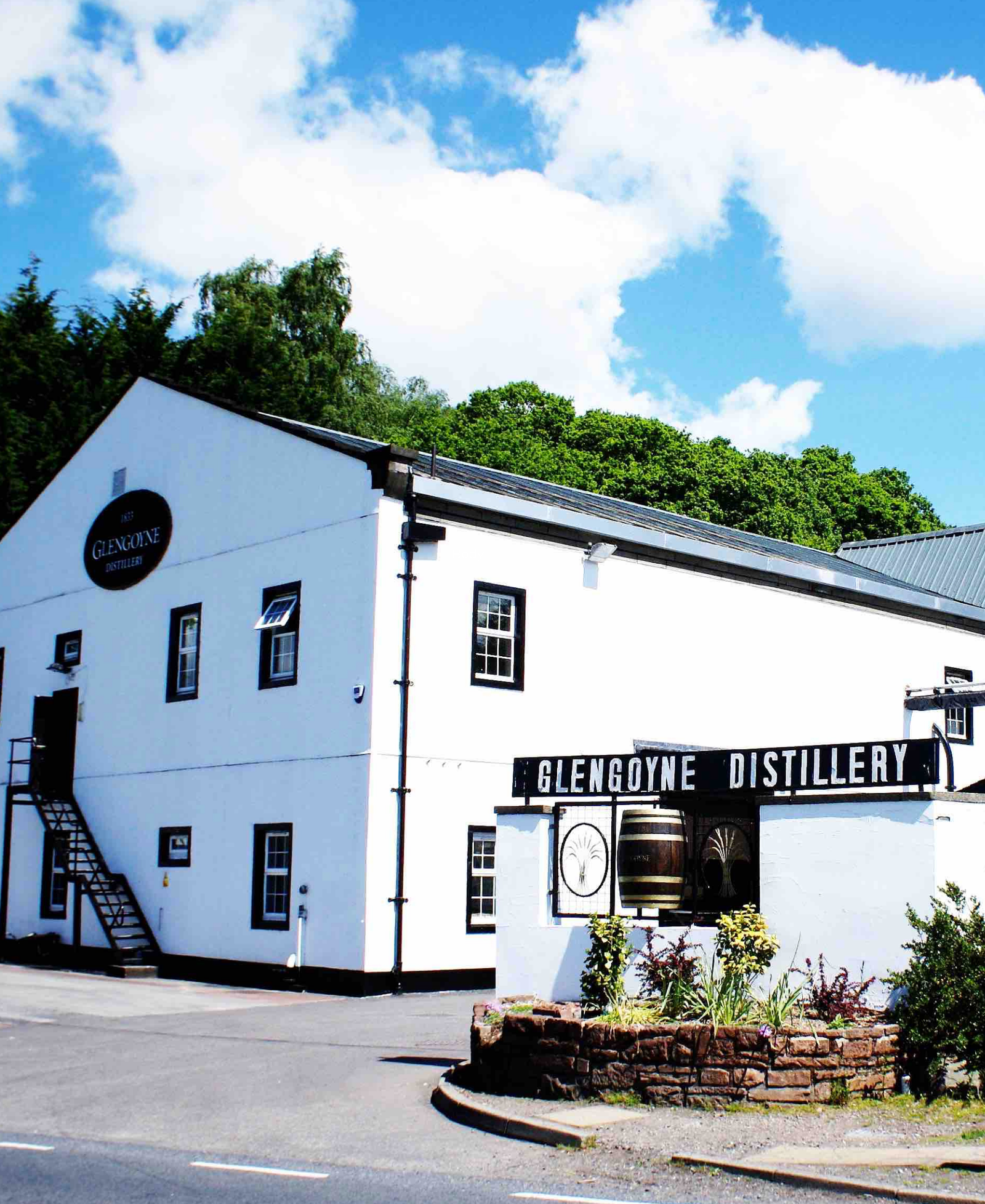 Glengoyne Distillery Glengoyne Distillery, a whitewashed Scottish whisky distillery building with signage and a whisky barrel display, set against wooded hills