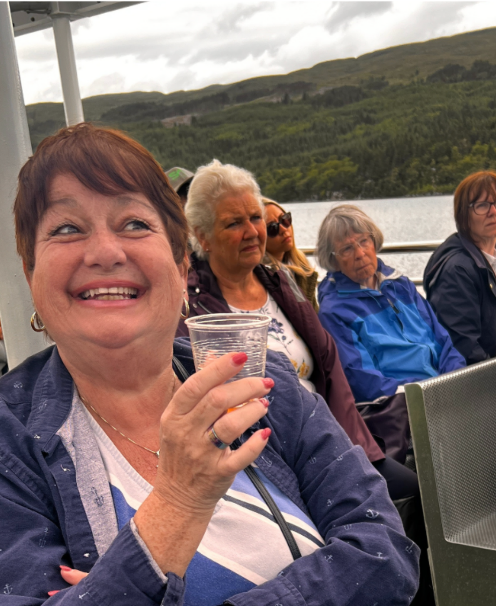 Guest on Loch Ness A smiling guest holding a drink aboard a boat tour on Loch Ness, with other passengers and wooded hills behind