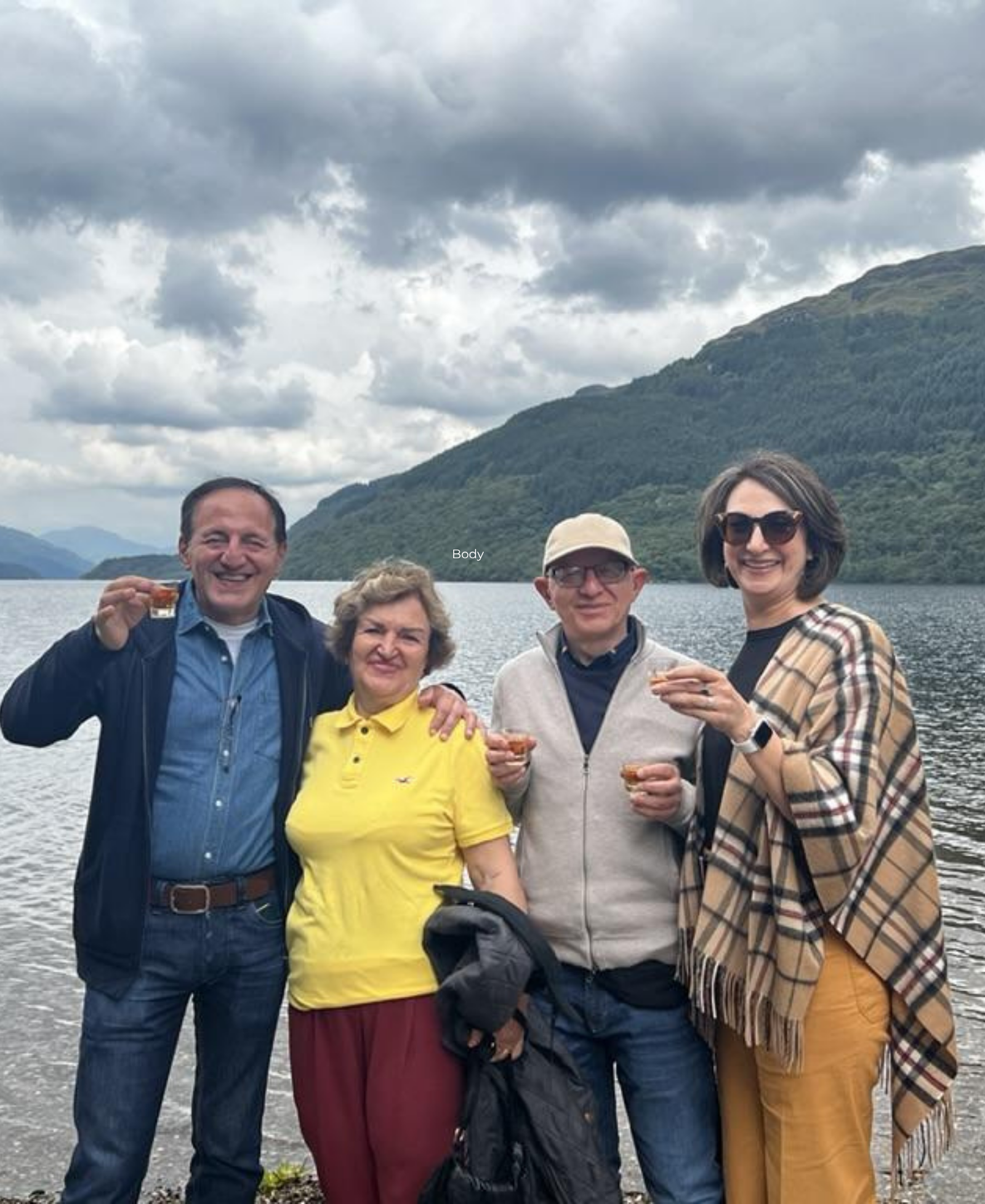 Guests at Loch Lomond Four guests raising small whisky glasses in a toast beside Loch Lomond, with mountains under a cloudy sky