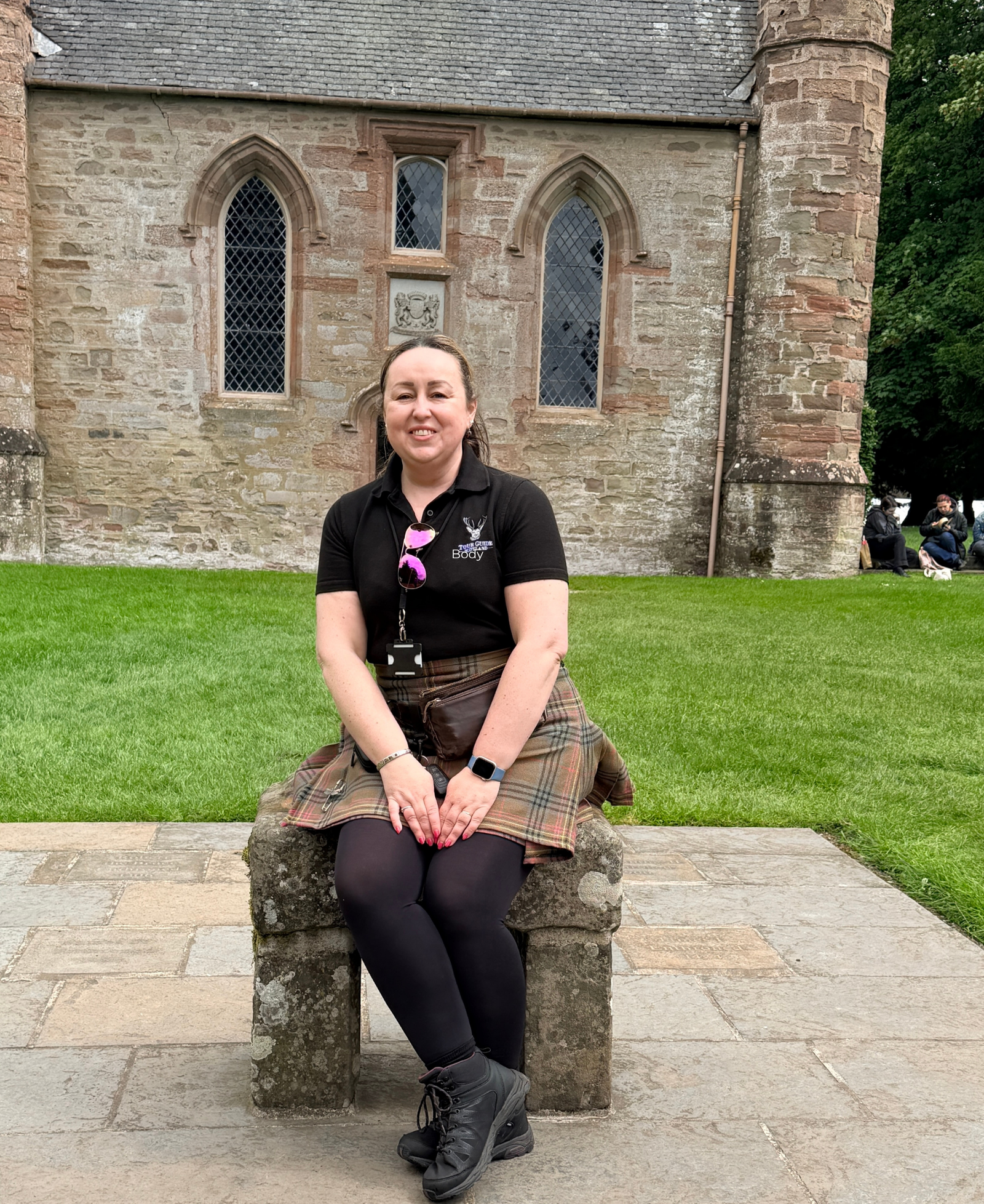 Lesley on the Stone of Destiny A tartan-clad tour guide sitting on the replica Stone of Destiny outside a chapel at Scone Palace