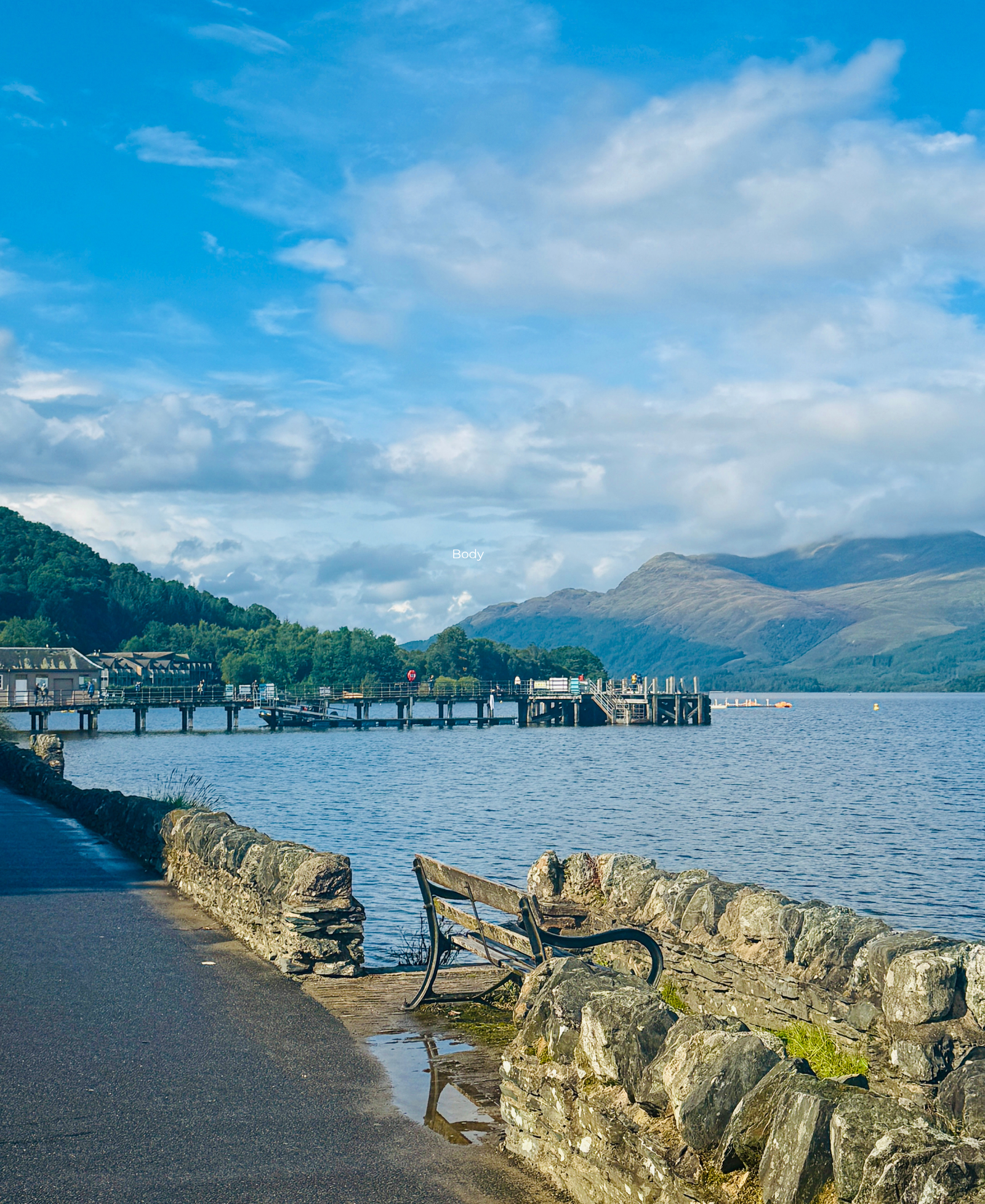 A wooden pier stretching into Loch Lomond, seen past a stone wall and bench, with mountains in the background