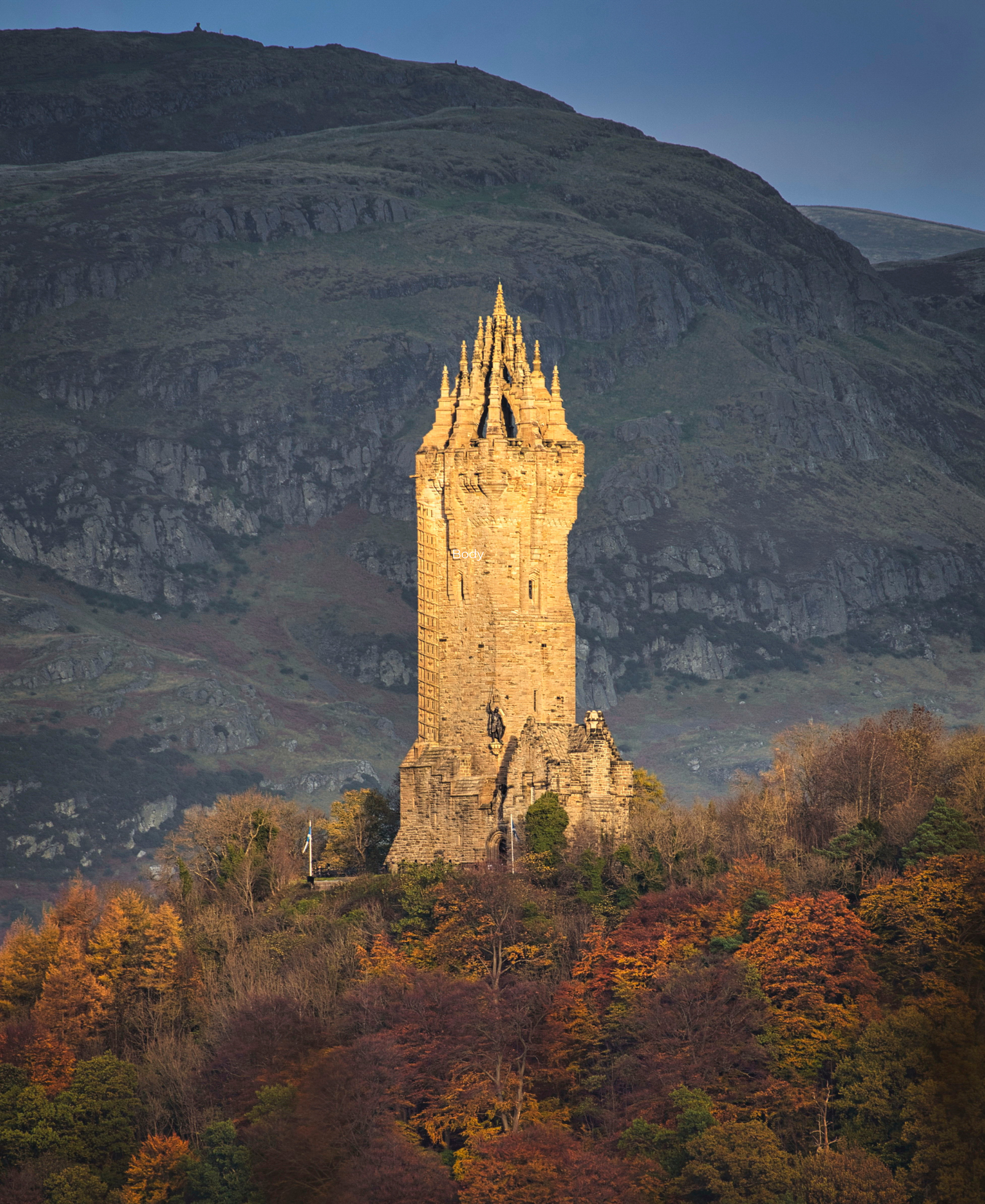 National Wallace Monument The National Wallace Monument tower lit by golden light, rising above autumn woodland with a dark hillside behind