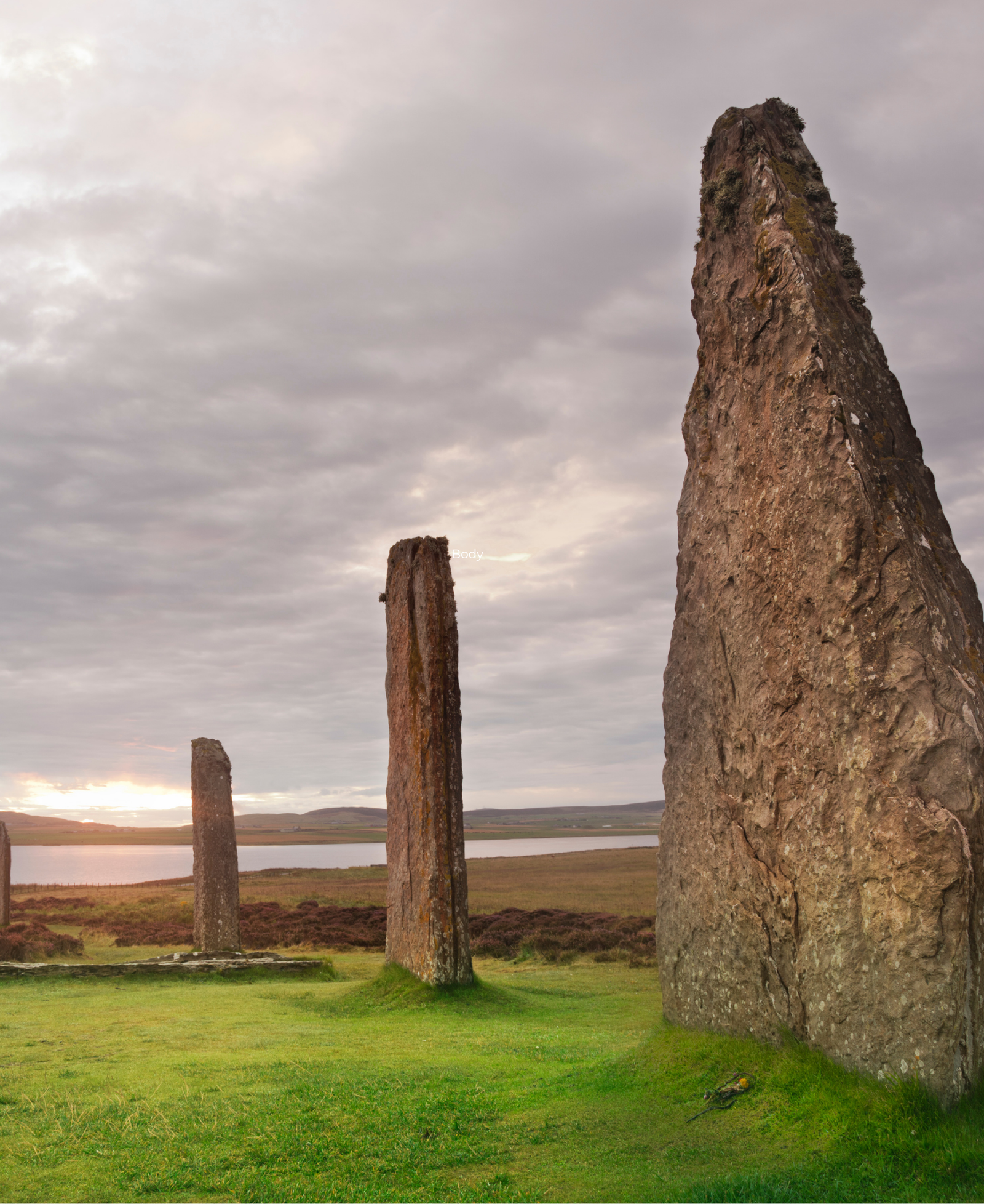 Ancient standing stones rising from green grass under a cloudy sky, with a loch and hills in the background