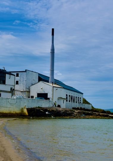Bowmore whisky distillery on the shoreline of Islay, with its tall chimney and 'Bowmore' sign visible above the beach