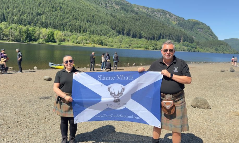 Meet James & Lesley Meet James & Lesley - tour guides in kilts holding a Tour Guide Scotland banner on a lochside beach, with forested hills behind