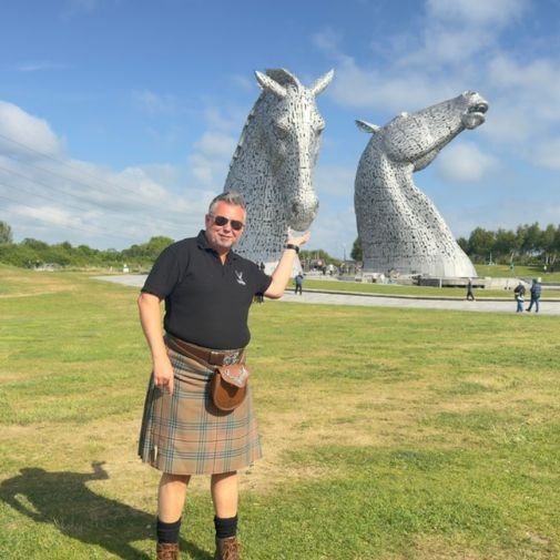 James at the Kelpies James from Tour Guide Scotland posing playfully with the Kelpies sculptures in Falkirk, as if feeding one of the horse heads
