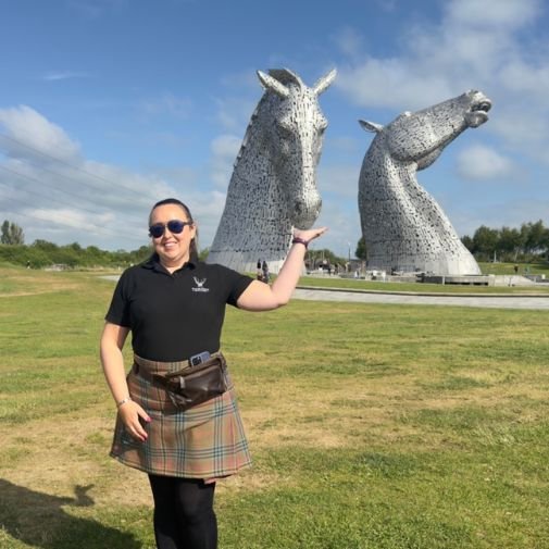 Lesley at the Kelpies Lesley from Tour Guide Scotland posing playfully with the Kelpies sculptures in Falkirk, as if feeding one of the horse heads