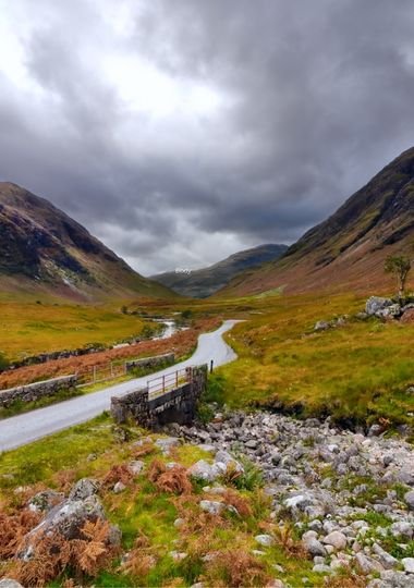 A single-track road winding through a Scottish Highland glen, with a small stone bridge and moody storm clouds overhead