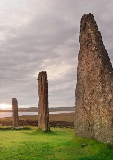 Ancient standing stones rising from green grass under a cloudy sky, with a loch and hills in the background